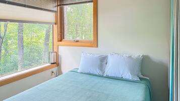 Ein Schlafzimmer mit einem Bett, einem Fenster mit Jalousien und Blick auf Bäume.