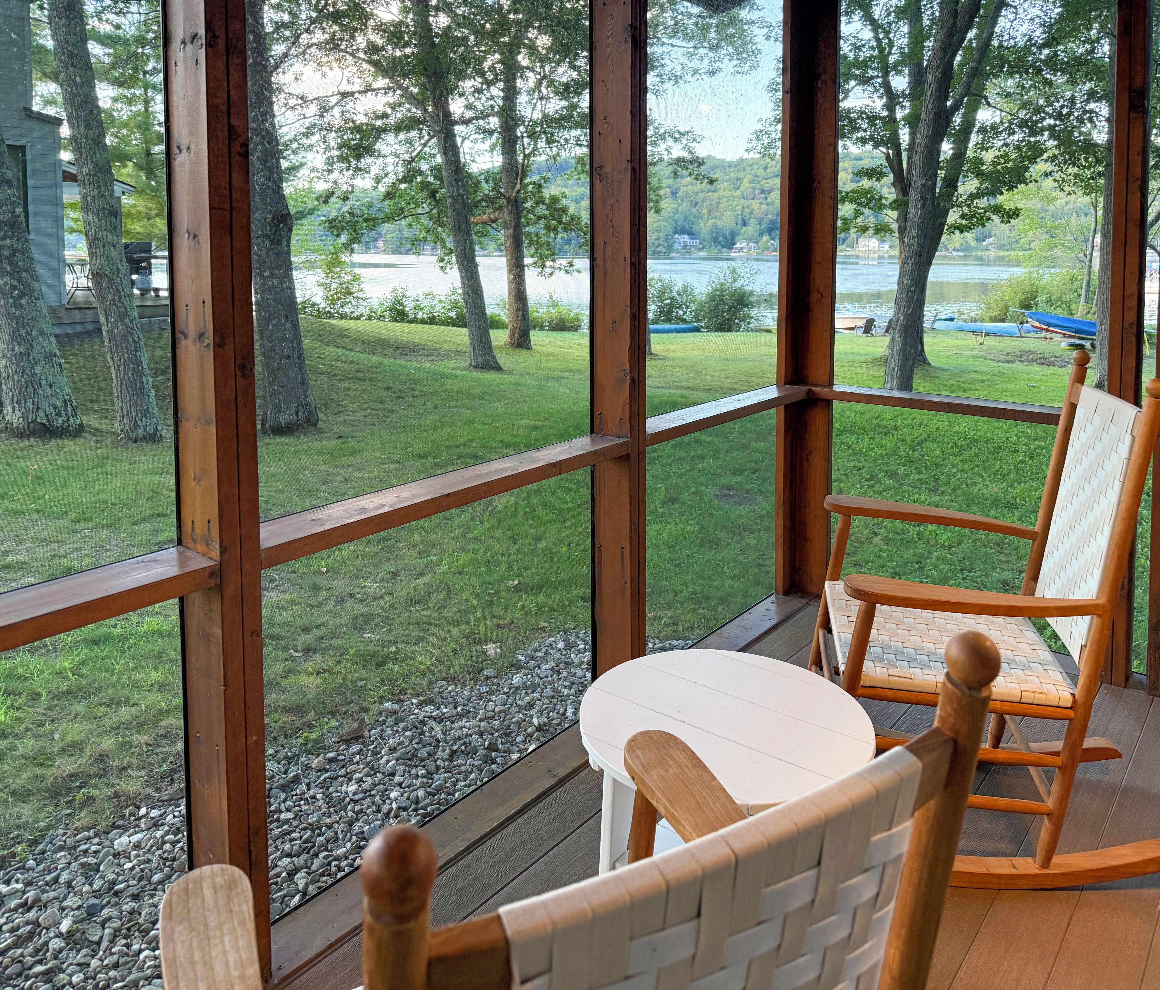 Eine Holzterrasse mit Tisch und Stühlen, mit Blick auf eine Rasenfläche mit Bäumen und einem Gewässer.