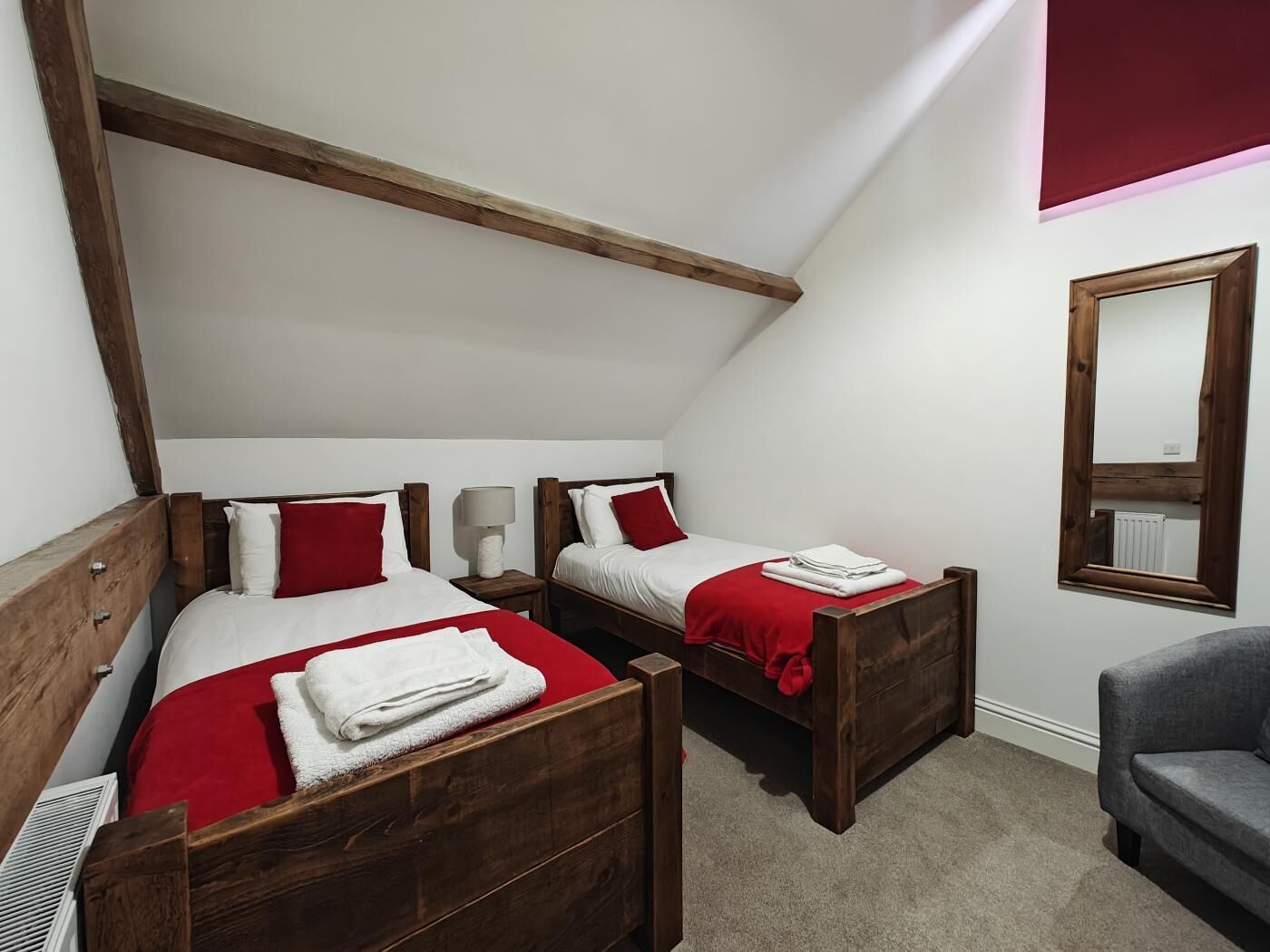 A bedroom with two wooden beds, each with a red blanket and white pillows, a grey sofa, and a mirror on the wall.