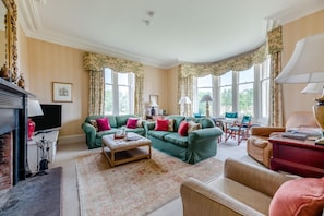 A spacious living room with a fireplace, green sofas, a coffee table, and large windows with curtains. - 5 Bed in Cornhill-on-Tweed (oc-s45856) (Cornhill-on-Tweed)