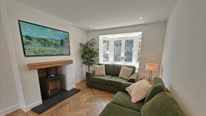 A living room with a green sofa, a fireplace, a painting, and a window with a view of greenery. - 4 Bed in Seahouses (oc-h45933) (Seahouses)