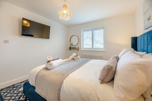 A neatly made bed with a blue headboard, a television mounted on the wall, a window with blinds, and a patterned rug. - 4 Bed in Seahouses (oc-h45933) (Seahouses)