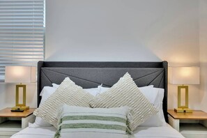 Ein ordentlich bezogenes Bett mit grauem Kopfteil, weißen Kissen und einer grün-weißen Streifenauflage. Auf beiden Seiten des Bettes stehen Nachttische mit goldfarbenen Lampen.