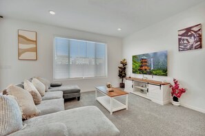 Ein modernes Wohnzimmer mit grauem Sofa, einem Couchtisch, einem Fernseher auf einem weißen Schrank und einem großen Fenster mit Jalousien.
