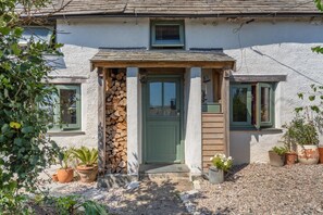 Ein Haus mit grüner Tür, einer kleinen Veranda und einem Holzstapel. Vor dem Eingang stehen Blumentöpfe.