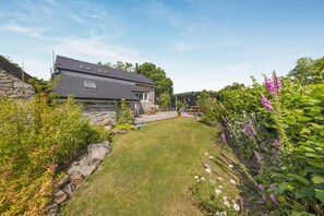 Ein Steinhaus mit schrägem Dach, einem kleinen Rasen und verschiedenen Pflanzen und Blumen.