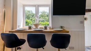 A dining area with a table set for two, a TV mounted on the wall, and a window with a view of greenery. - 2 Bed in Llanwrthwl (oc-b45319) (Llanwrthwl)