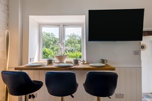 Ein Essbereich mit einem Tisch für zwei Personen, einem an der Wand befestigten Fernseher und einem Fenster mit Blick auf Grünflächen.
