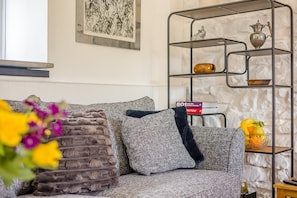 Una sala de estar con un sofá gris, una estantería metálica con objetos decorativos y una obra de arte enmarcada en la pared.