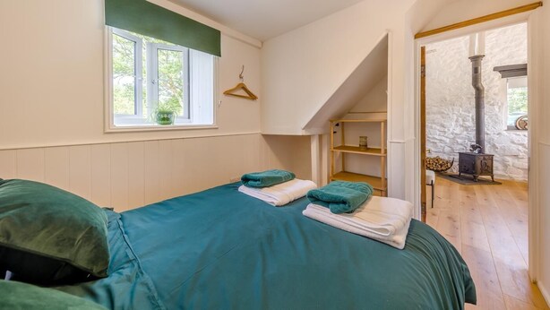 A bedroom with a bed, a window, a hanging coat rack, a wooden shelf, and a stone wall with a fireplace. - 2 Bed in Llanwrthwl (oc-b45319) (Llanwrthwl)