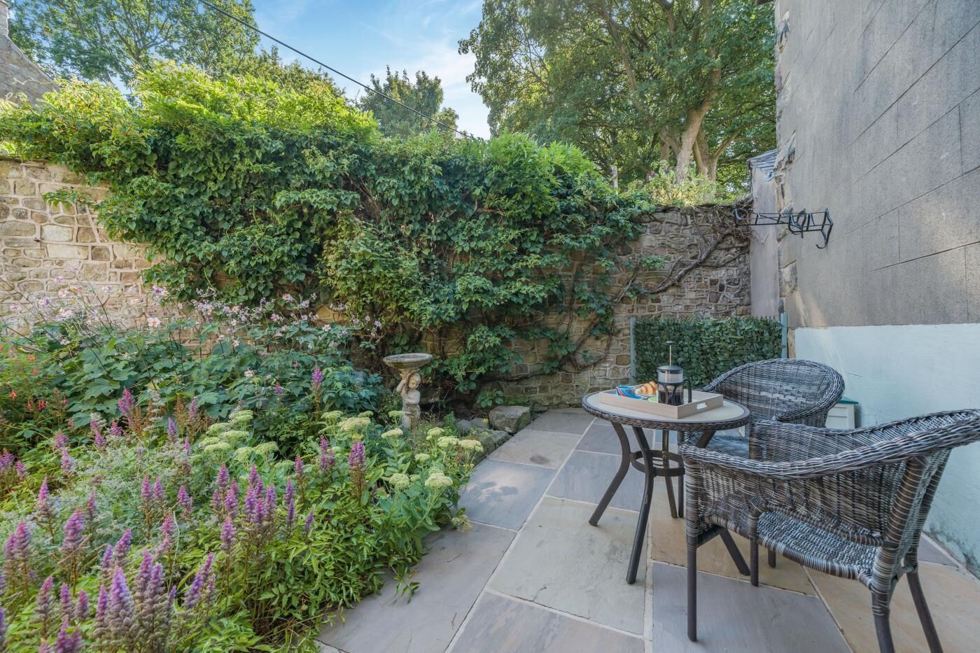 Un patio con suelo de piedra, una mesa con sillas y un muro de piedra cubierto de plantas trepadoras.