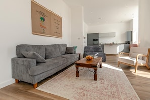 A living room with a grey sofa, a wooden coffee table, and a kitchen in the background. - 3 Bed in Ilfracombe (oc-b45837) (Ilfracombe)