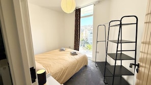 A hotel room with a bed, a window with a view of buildings, a black metal shelf unit, and a lamp on a bedside table. - 3 Bed in Ilfracombe (oc-b45837) (Ilfracombe)