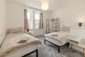 A modern bedroom with two beds, a bedside table, a hanging light fixture, and a window with curtains. - 3 Bed in Ilfracombe (oc-b45837) (Ilfracombe)
