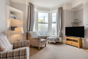 A living room with a sofa, armchair, ottoman, a flat-screen TV on a wooden stand, a lamp, and a bookshelf with a laptop. - 4 Bed in Brixham (oc-a45940) (Brixham)