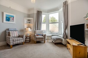 A cozy living room with two armchairs, a small table, a flat-screen TV, and a bay window with curtains. - 4 Bed in Brixham (oc-a45940) (Brixham)