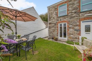 A patio with a table and chairs, a white umbrella, and a stone building with a window and door. - 3 Bed in Treknow (oc-p45966) (Treknow)