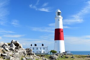 A lighthouse with a white and red color scheme, a white building with multiple windows in the background, and a rocky shoreline. - 2 Bed in Weymouth (oc-m45968) (Weymouth)