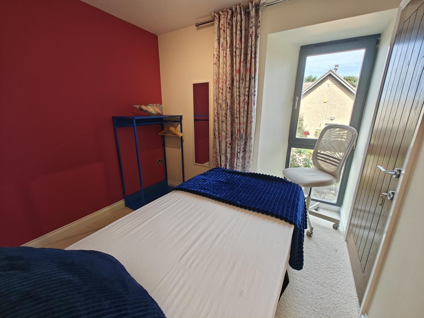 A bedroom with a bed, a desk, and a chair. There is a window with curtains and a view of the outside.