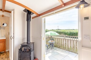 A room with a wood-burning stove, a wooden door, a small table with a chair, and a view of a vineyard through an open door. - 1 Bed in St Austell (oc-h45926) (High Street)