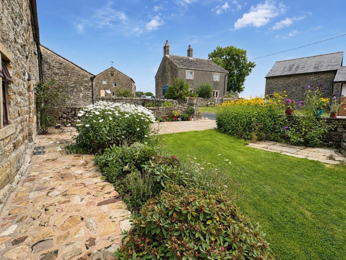 A stone building with a stone pathway leading to a grassy area with various plants and flowers.