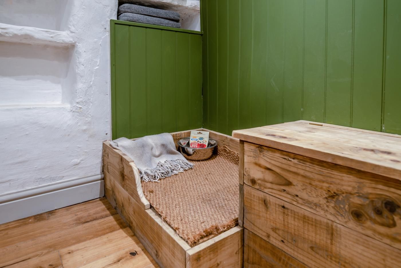 A wooden crate with a blanket and a bowl of dog food, a wooden dresser, and a green wall.