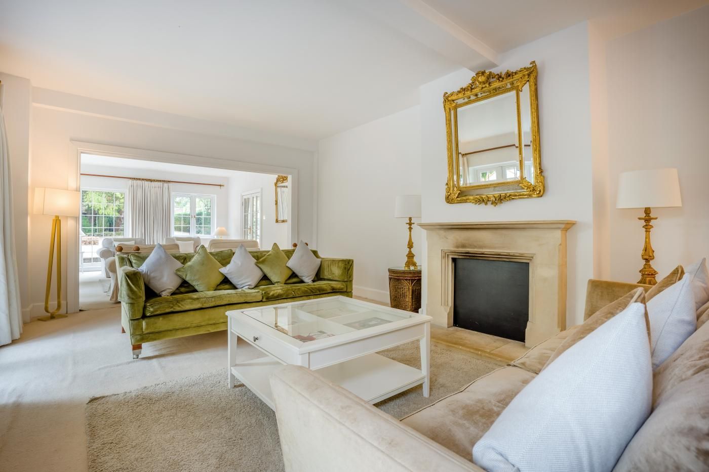 Una sala de estar con un sofá verde, sillones blancos, una mesa de centro de vidrio, una chimenea y un espejo grande.