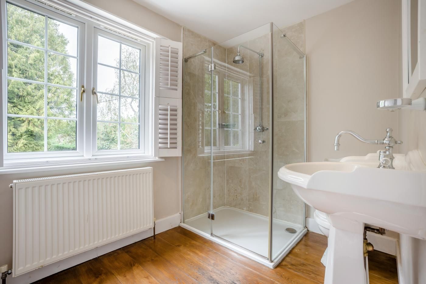 Een badkamer met een douchecabine van glas, een witte wastafel en een raam met zonwering.