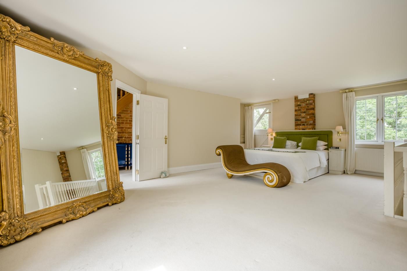 A spacious bedroom with a large mirror, a bed with green headboard, a chaise lounge, and a brick accent wall.