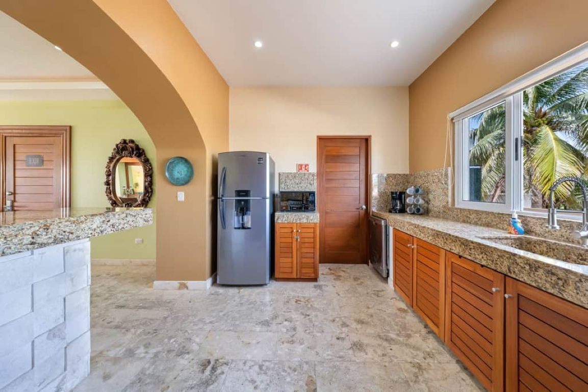Una cocina con piso de mármol, gabinetes de madera, un refrigerador de acero inoxidable y un ventanal con vista a palmeras.