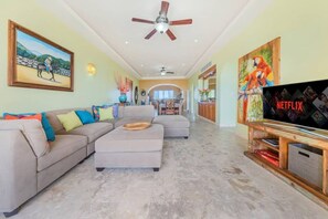 A living room with a sofa, ottoman, ceiling fan, artwork, and a television. - Luxury Oceanfront 2-Bedroom Condo at Isla 33 (Isla Mujeres)