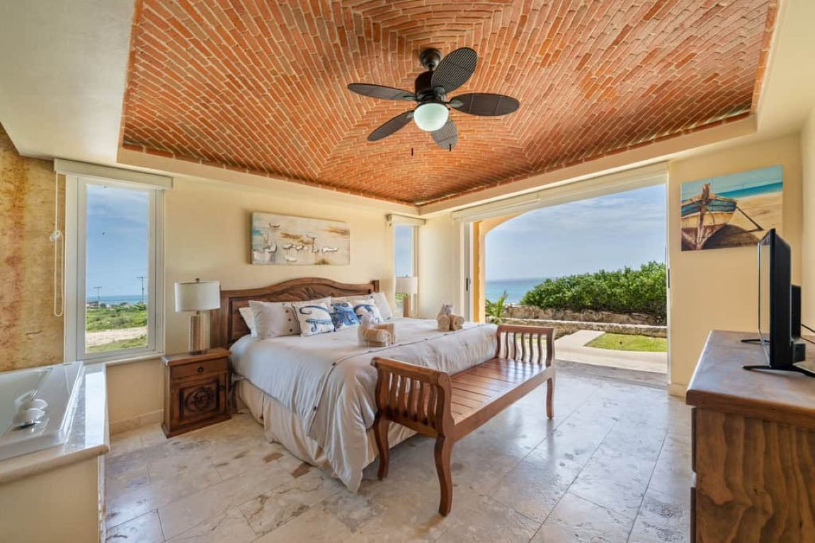 Een slaapkamer met een bed, een houten bankje, een plafondventilator en uitzicht op zee.