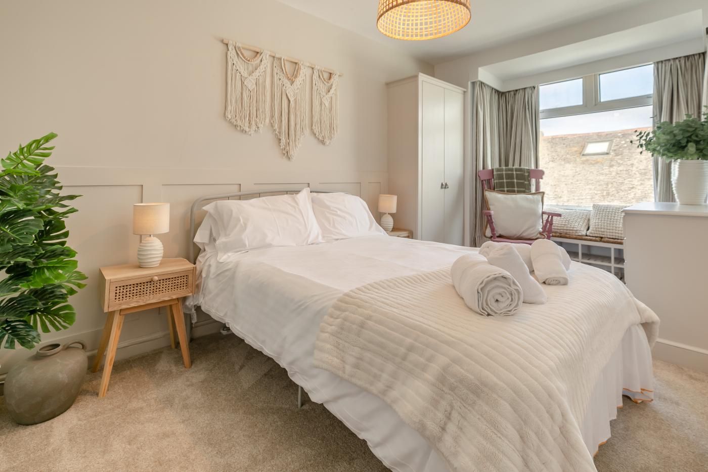 A neatly made bed with a white comforter and pillows, a bedside lamp, a macrame wall hanging, and a view of the outdoors through a window.