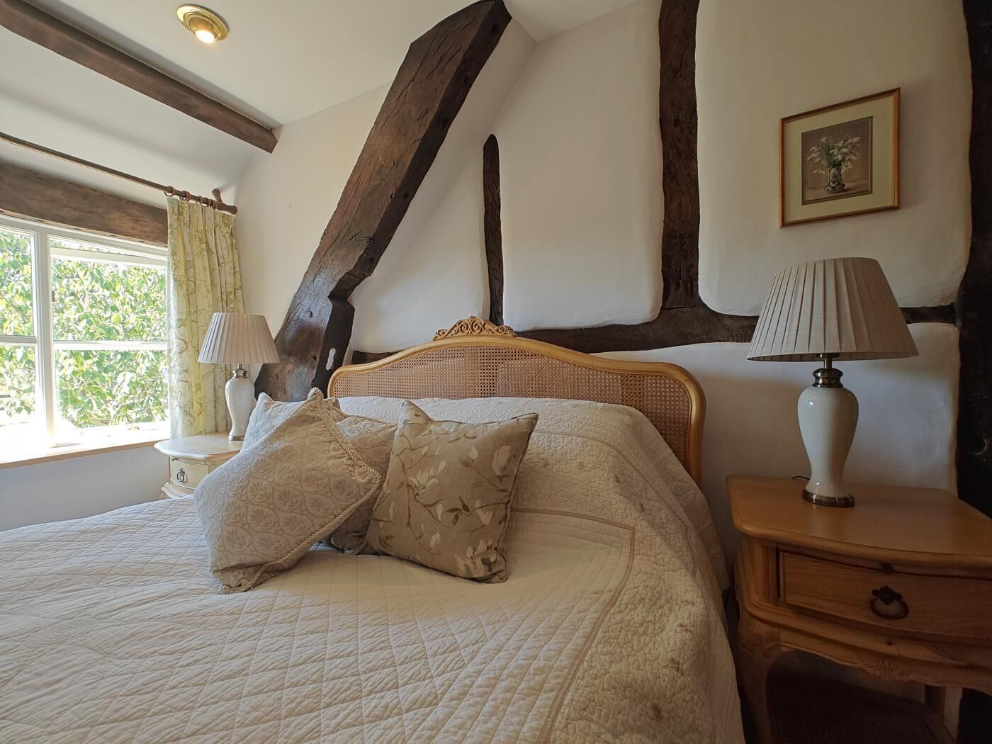 Een slaapkamer met een bed, een nachtkastje, een lamp, een raam met gordijnen en een lijst aan de muur.
