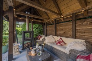 A covered outdoor seating area with a wicker sofa, cushions, a table with food and drinks, and a fireplace. - 1 Bed in Botus Fleming (oc-f45947) (Botusfleming)