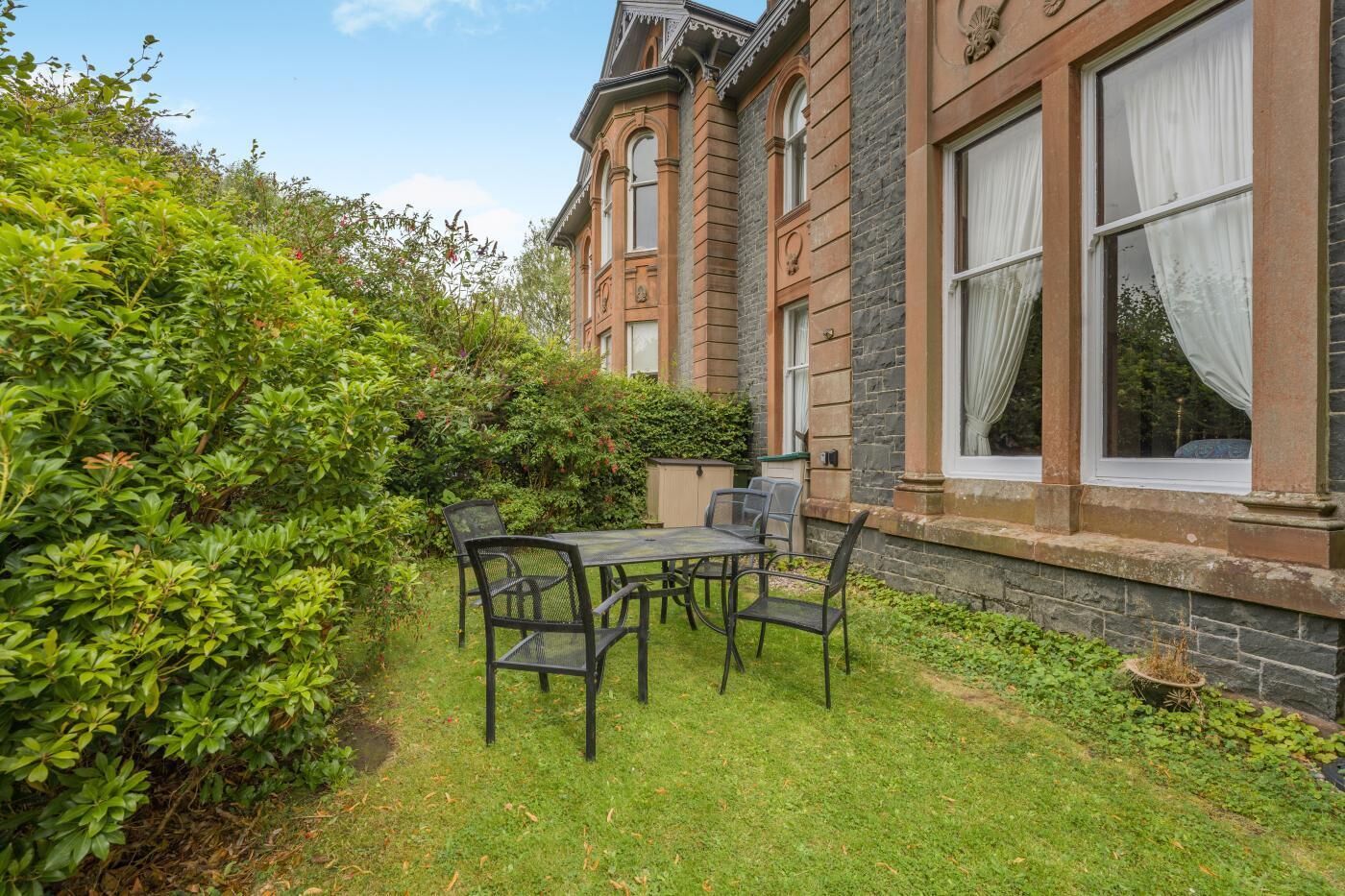 A traditional building with a stone facade, a garden with a table and chairs, and a clear sky.