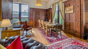 Een kamer met een houten eettafel, stoelen, een leren bank, een lamp en een groot raam met jaloezieën.