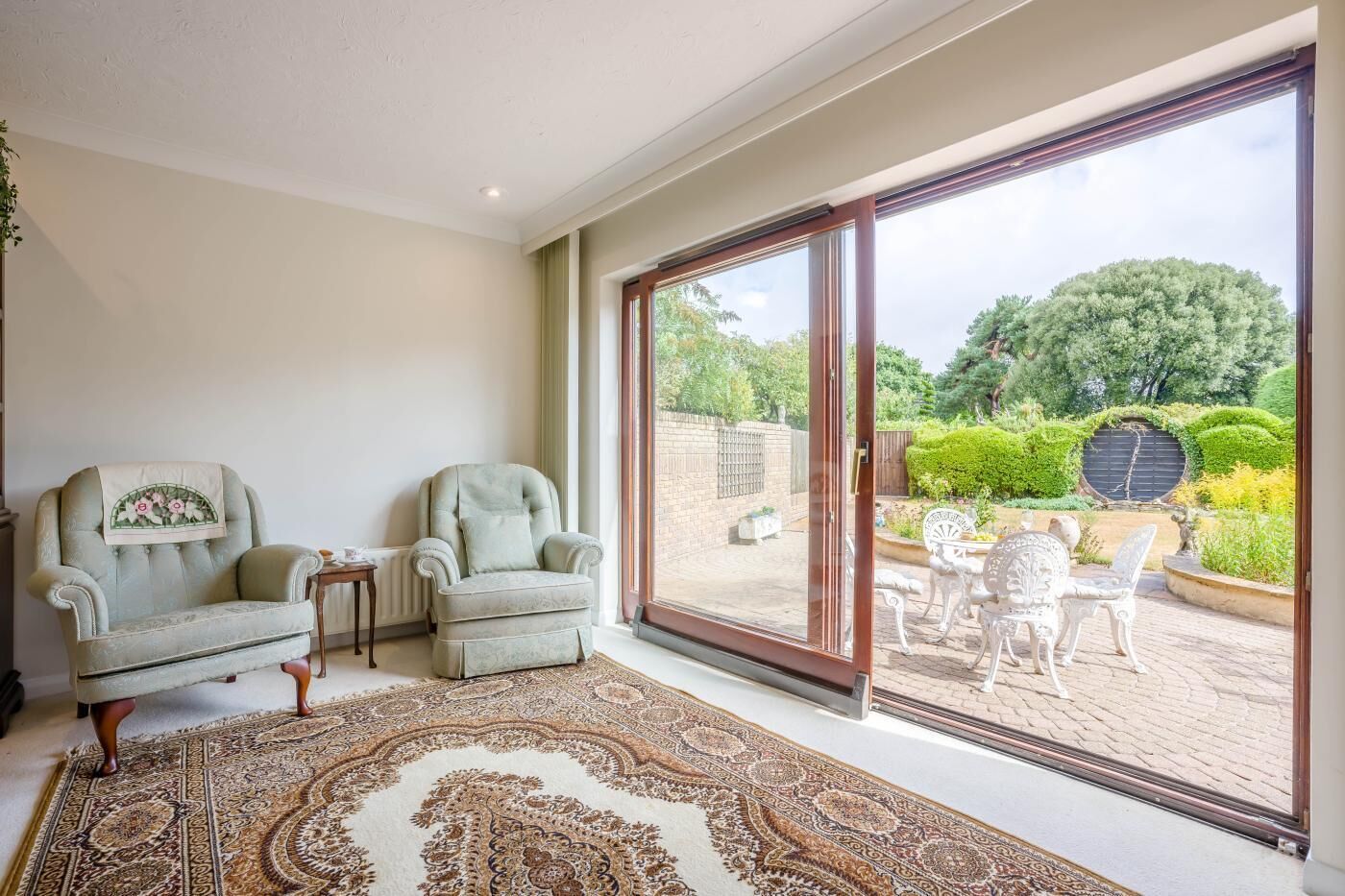 Ein Zimmer mit zwei Sesseln, einem gemusterten Teppich und Blick auf einen Garten durch eine Schiebetür.