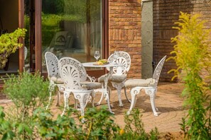Eine Terrasse mit weißer Gartenmöblierung, einem Tisch und Stühlen, vor einer Ziegelwand mit einer Glastür, die in einen Garten führt.