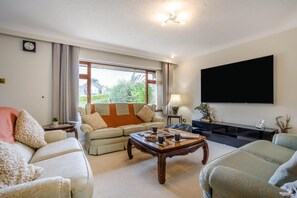 Ein Wohnzimmer mit einer Couch, Sesseln, einem Couchtisch, einem Fernseher und einem Fenster mit Vorhängen.