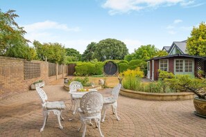 A patio with a brick floor, white ornate chairs, a table with a wine glass, and a small building with a red door. - 3 Bed in Christchurch (oc-m45917) (Christchurch)