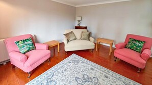 A living room with two pink armchairs, a beige sofa, a patterned rug, wooden side tables, and a wall-mounted lamp. - 2 Bed in Hoxne (oc-b45969) (Hoxne)