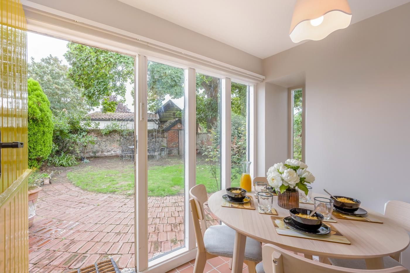 A dining area with a table set for two, a vase of white flowers, and a view of a garden through large windows.