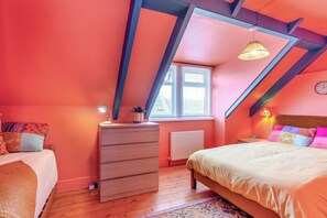 Ein Schlafzimmer mit schrägem Dach, einer hölzernen Kommode, einem Bett mit farbigem Kopfteil und einem Fenster mit Jalousien.