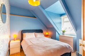 Ein Schlafzimmer mit schrägem Dach, einem Bett mit karierter Bettwäsche, Nachttischlampen, einer Kommode und einem Fenster mit Jalousien.