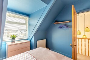 A bedroom with a sloped ceiling, a radiator, a bed with a checkered blanket, a window with blinds, a potted plant, and a wooden door with a light fixture. - 4 Bed in Newquay (oc-t45967) (Newquay)