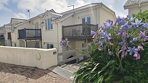 Une rangée de maisons à deux étages, dotées de balcons, bordée d’une allée pavée et d’un jardin où poussent des fleurs violettes.