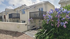 A row of two-story houses with balconies, a paved walkway, and a garden with purple flowers. - 2 Bed in Newquay (oc-t45990) (Newquay)
