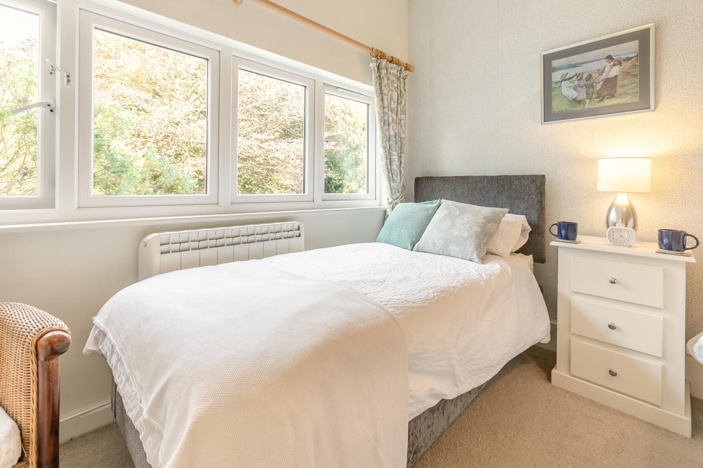 A bedroom with a bed, a bedside table with a lamp, a picture on the wall, and a window with curtains.