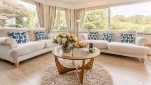 Ein modernes Wohnzimmer mit einem Glastisch, zwei weißen Sofas mit blauen Kissen und Blick ins Grüne.